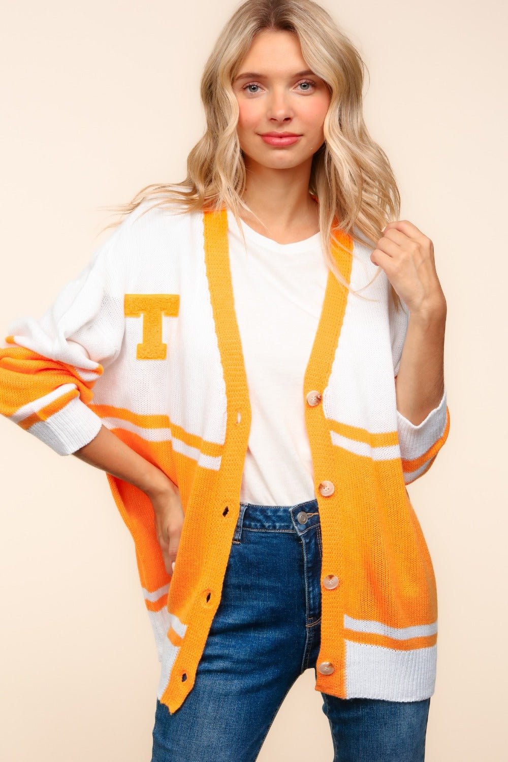 A person wearing the Haptics V Neck Button Down Letter Patch Cardigan, oversized with white and orange stripes and a yellow "T" patch, paired with blue jeans, smiles while lifting their hands to their head.