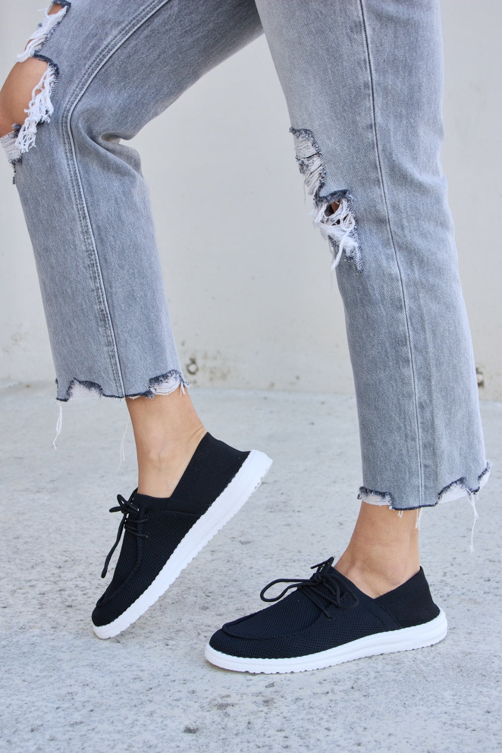 A person wearing ripped jeans and Forever Link Flat Round Toe Lace-Up Sneakers with white soles stands on a concrete surface.