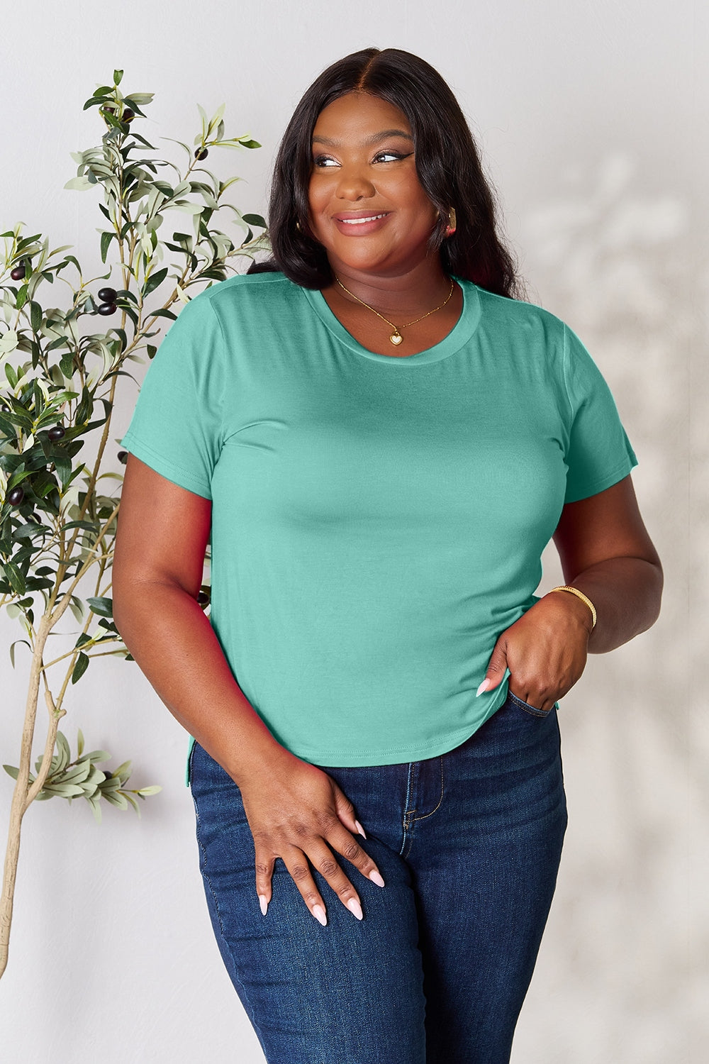 A person with wavy hair, wearing the Basic Bae Full Size Round Neck Short Sleeve T-Shirt in bright pink and beige pants, stands near a plant, smiling and looking to the side. The outfit embodies a basic style and offers slightly stretchy comfort, ideal for those who value effortless elegance.