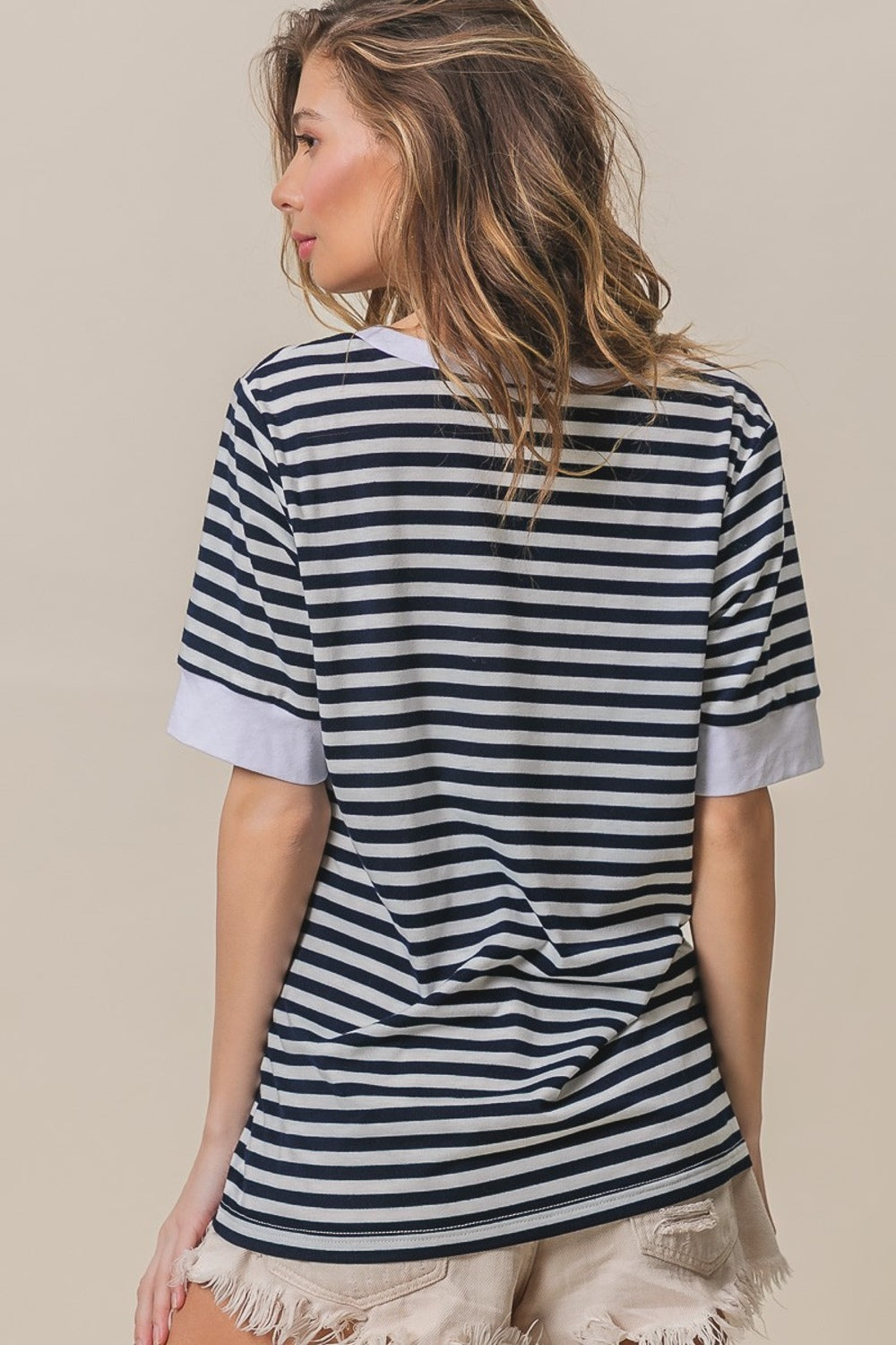 A person with long hair, wearing a BiBi Contrast Striped Notched Knit Top in navy and white and beige shorts, is posing with one hand in their hair, smiling against a beige background.