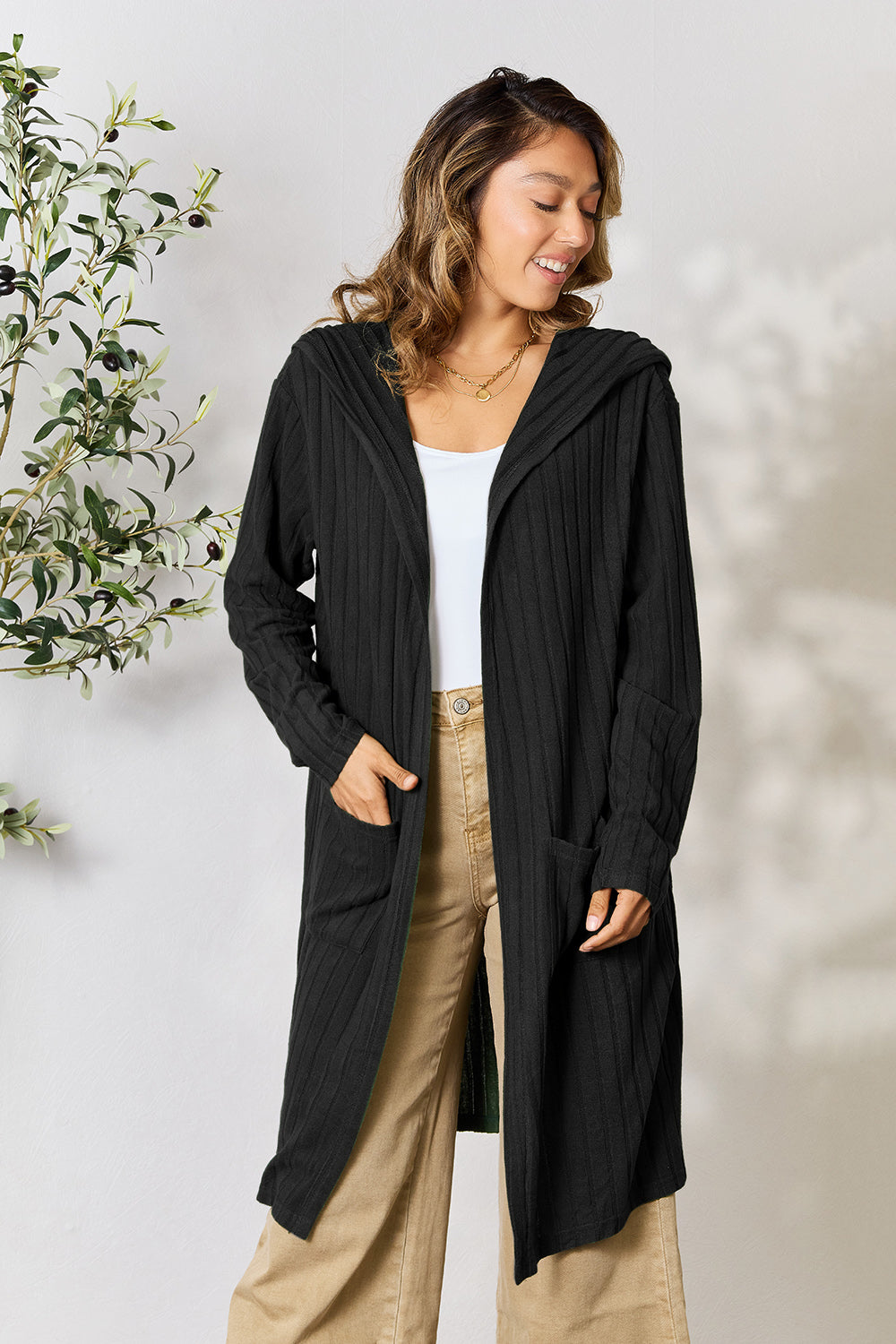 A woman wearing the Basic Bae Full Size Hooded Sweater Cardigan in black, paired with a white top and khaki pants, stands in front of a light background accented by a leafy plant on her left. With one hand tucked into her pocket, she gazes to her right.