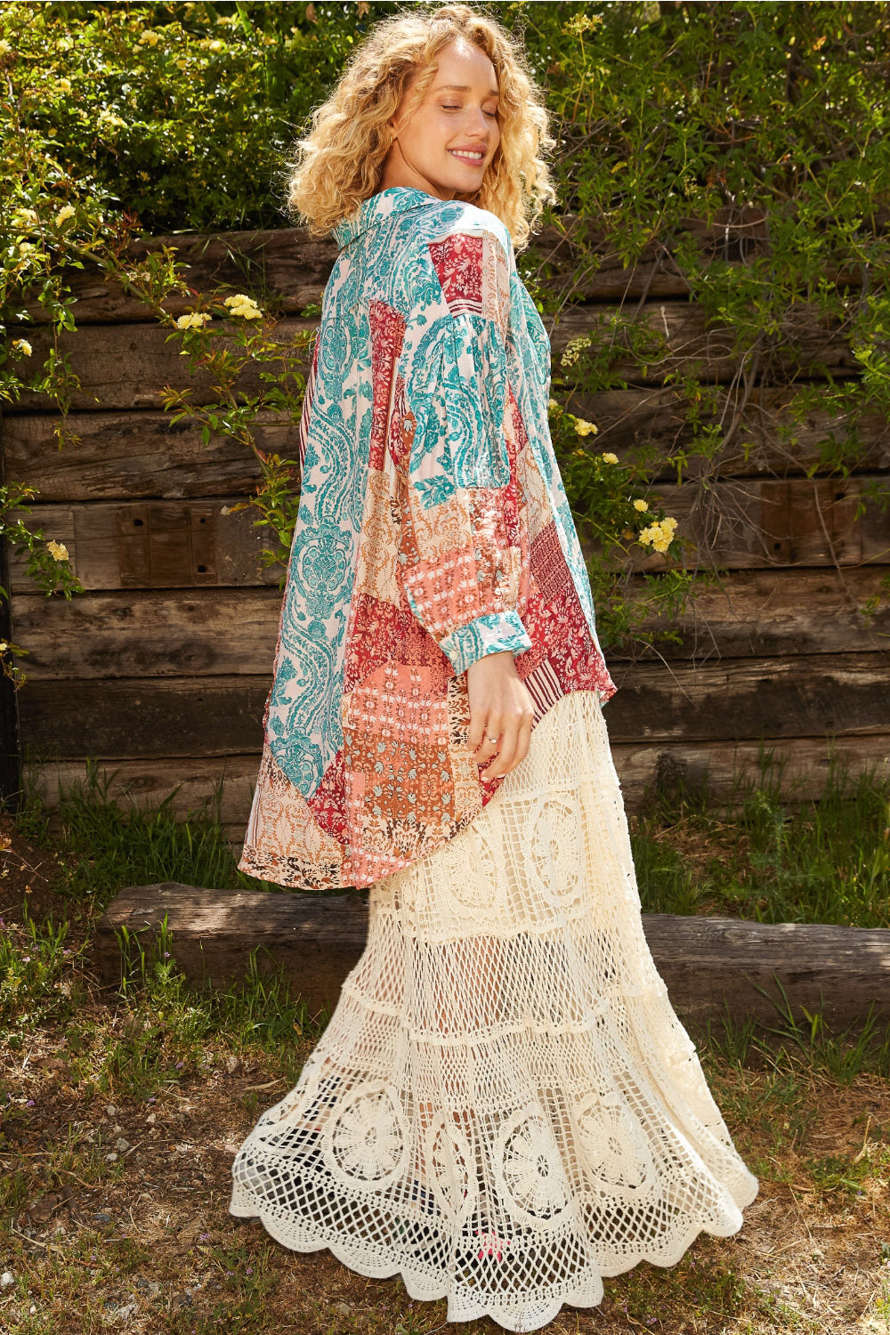 A person with curly hair models a colorful patchwork jacket over the POL Long Sleeve Printed Button Down Shirt and a cream-colored crochet skirt in an outdoor setting with greenery and a wooden fence.