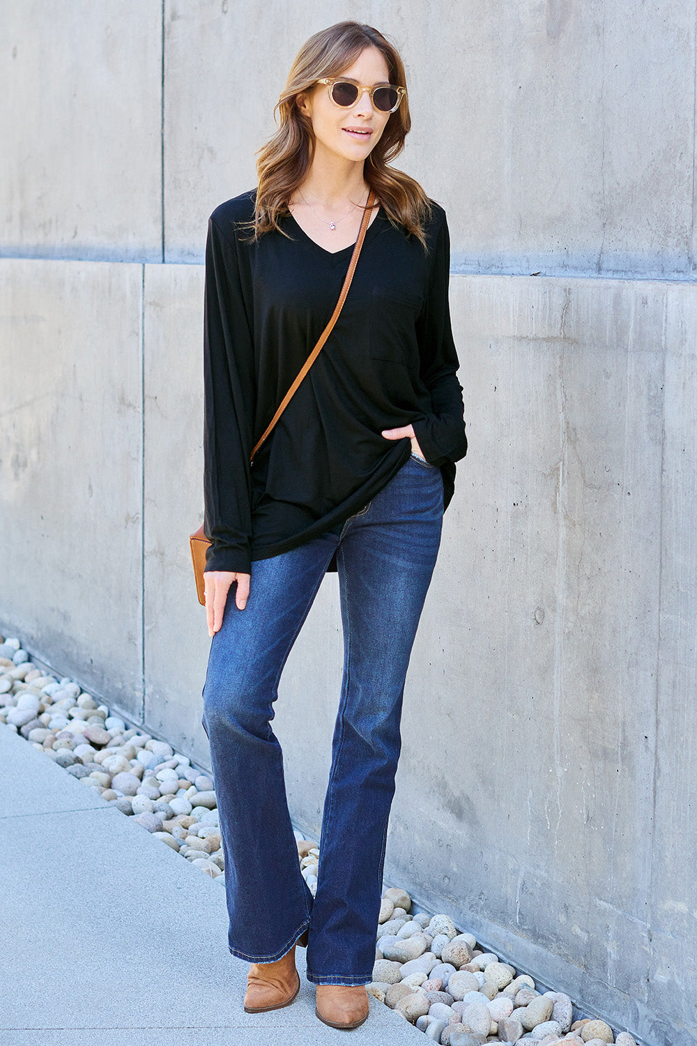 A woman with light brown hair stands against a concrete wall, wearing the Basic Bae Full Size V-Neck Long Sleeve Top in olive green and blue jeans, with a white backpack on her shoulder. Her outfit exudes basic style and is made from polyester, ensuring it’s both comfortable and suitable for machine wash cold. She is looking at the camera.