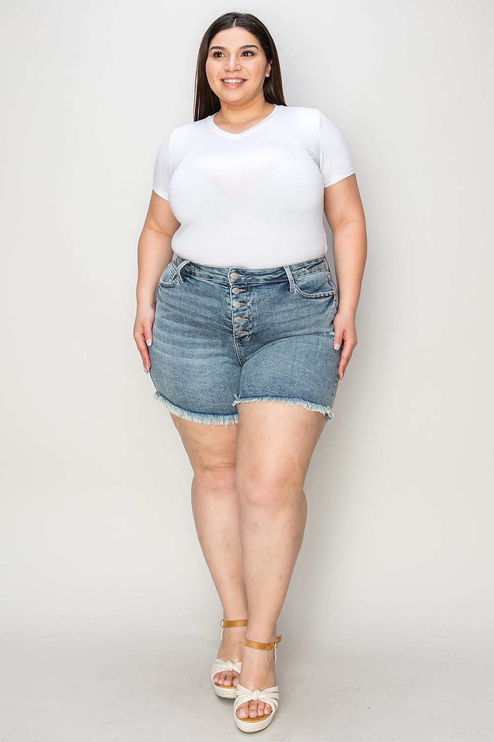 Wearing the Judy Blue Full Size Button Fly Raw Hem Denim Shorts and a white t-shirt, a person stands smiling in sandals against a plain background.