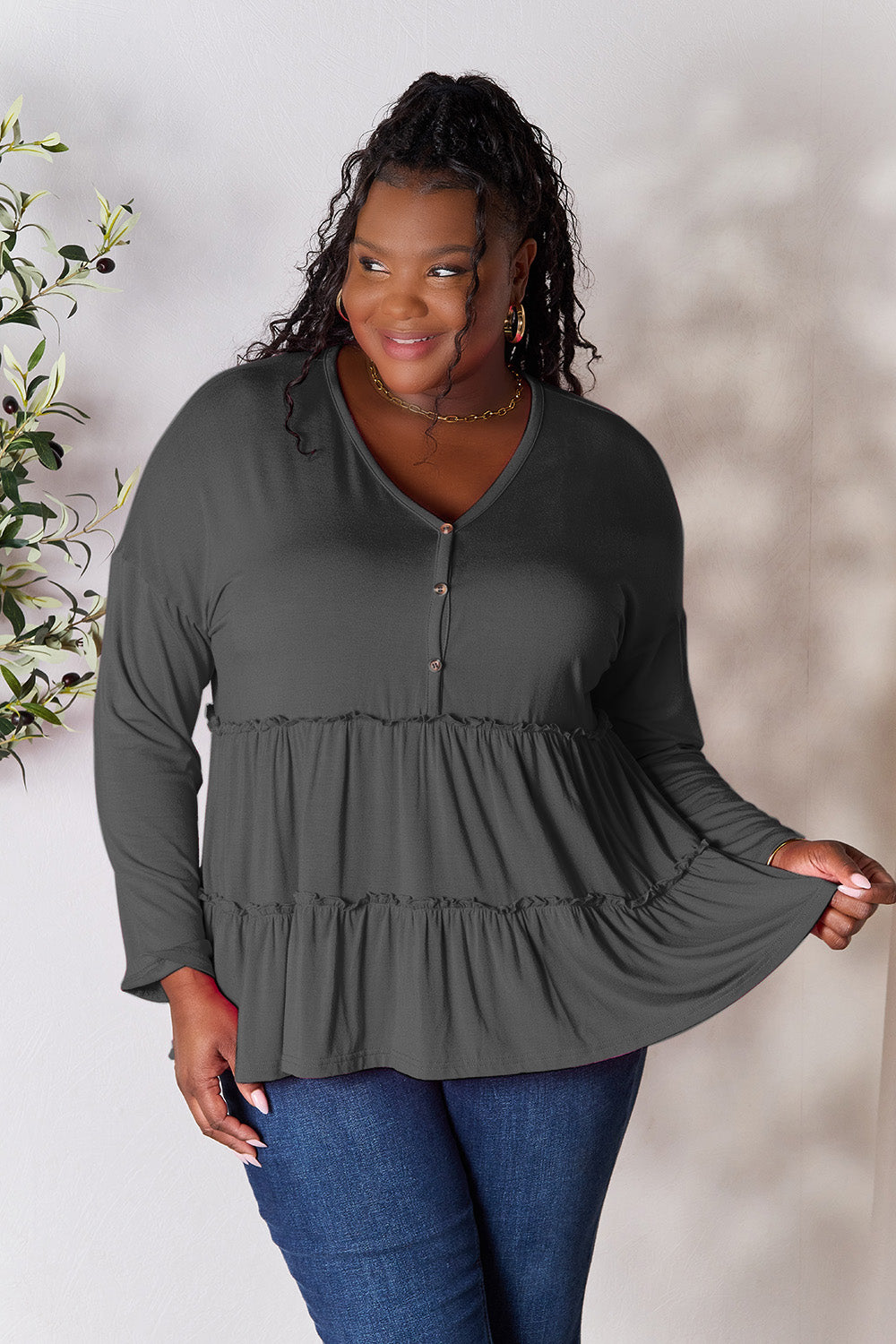 Person stands indoors near a plant, wearing the Double Take Half Button Long Sleeve Ruffle Hem Blouse in gray and blue jeans, smiling while looking to the side. The slightly stretchy fabric of the blouse adds comfort while its buttoned design offers a touch of elegance.