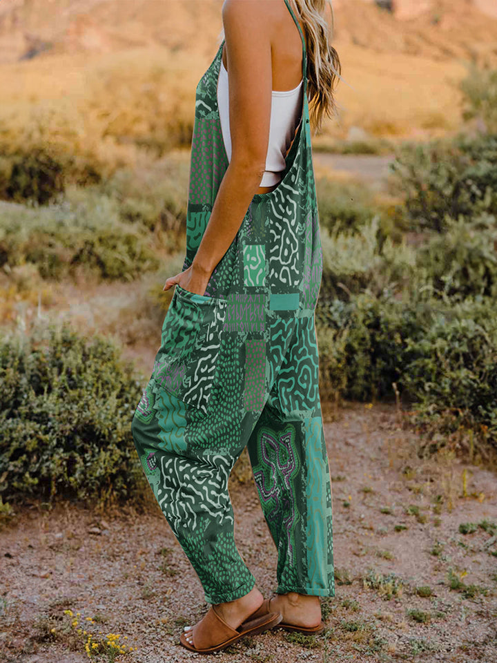 A person standing outdoors in front of rocky terrain, wearing a Full Size Printed V-Neck Sleeveless Jumpsuit and a hat.