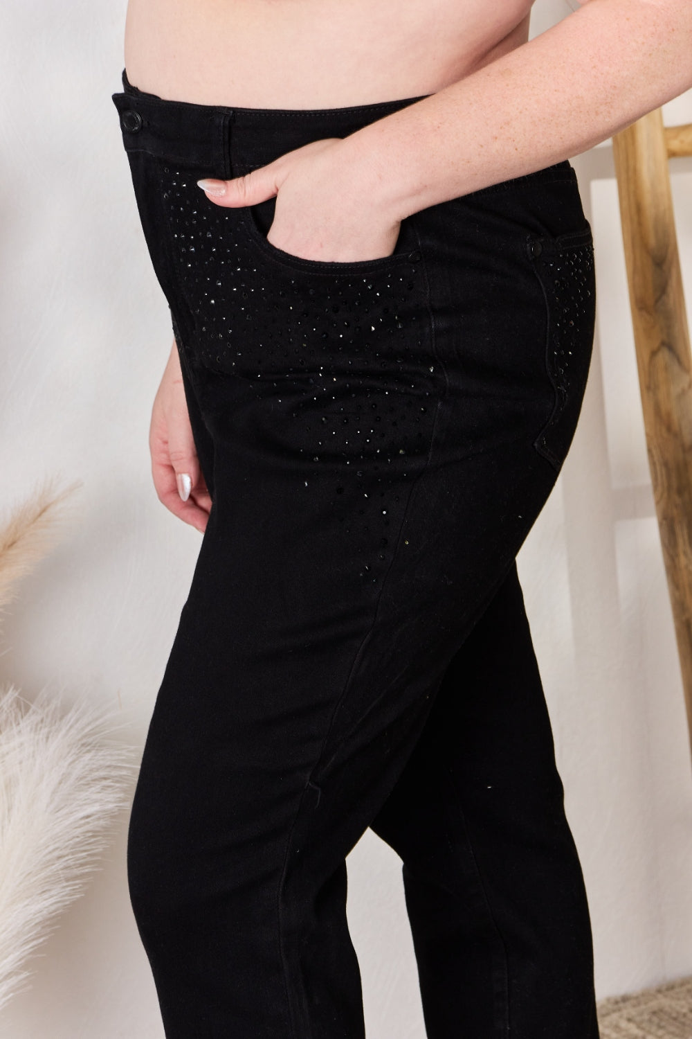 A person wearing slightly stretchy, black Judy Blue Full Size Rhinestone Embellished Slim Jeans and black shoes stands on a textured floor next to a white vase with brown and white ornamental grasses and a wooden ladder.