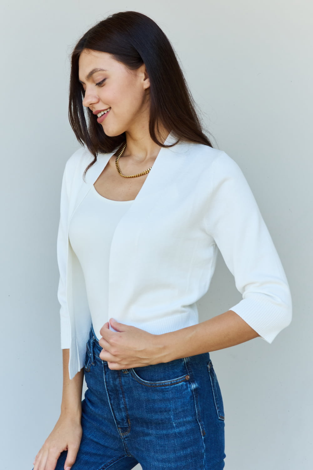 A stylish person wearing sunglasses, a Doublju My Favorite Full Size 3/4 Sleeve Cropped Cardigan in Ivory, and blue jeans poses in front of a plain background, effortlessly highlighting their versatile look.