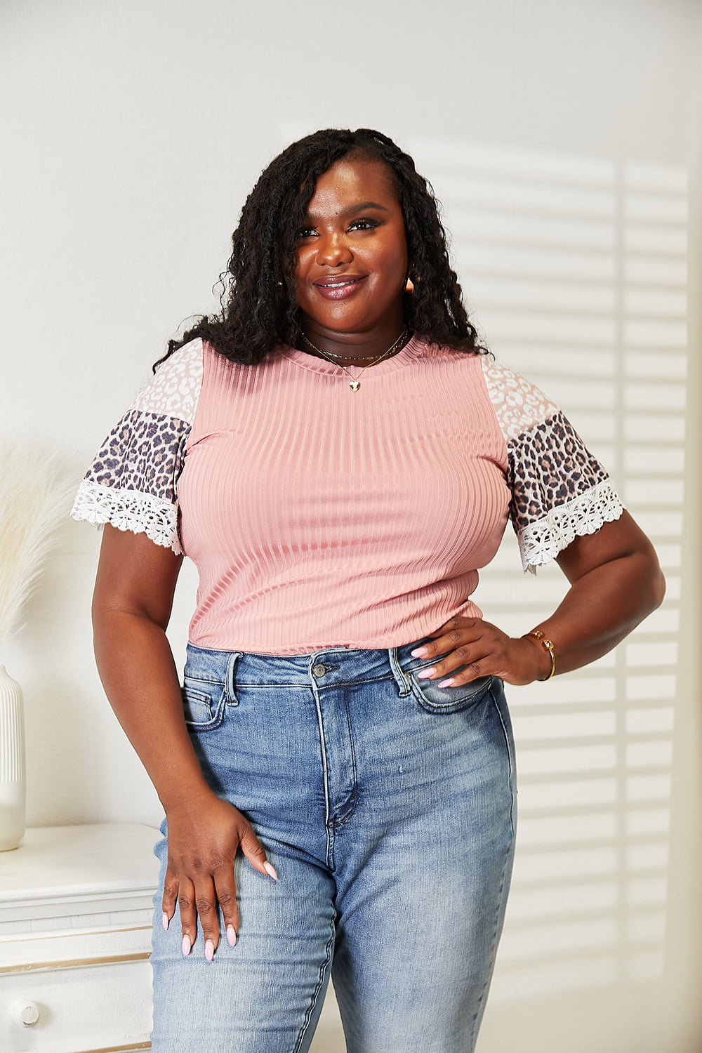 A woman with long hair wearing a trendy Double Take Leopard Lace Trim Ribbed Round Neck Top paired with blue jeans stands indoors smiling.