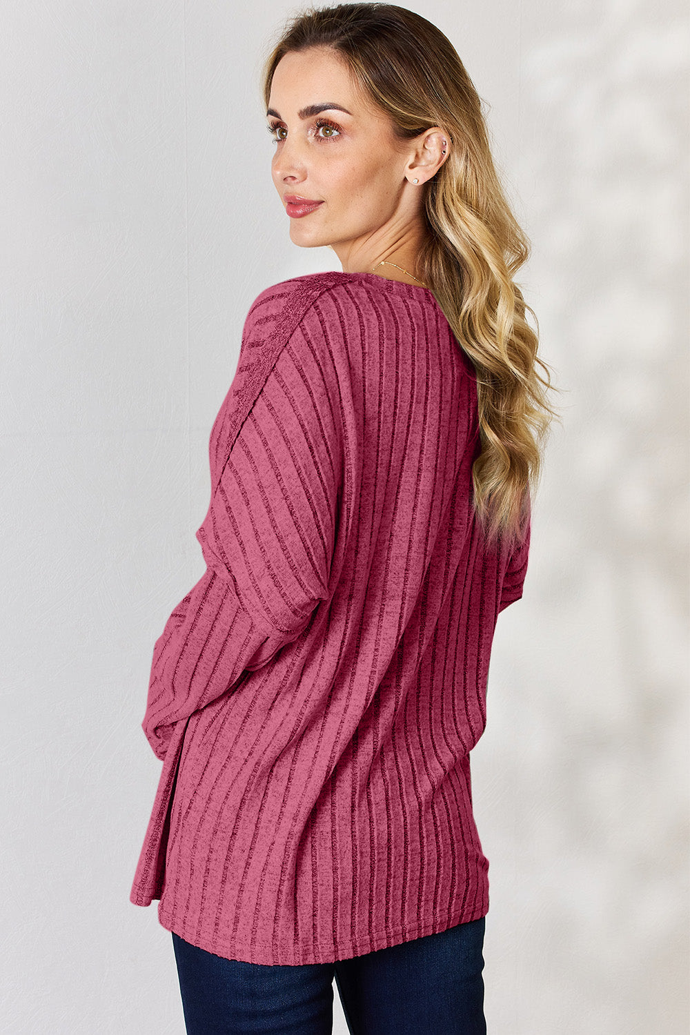 A woman with long, wavy blonde hair wears a slightly stretchy, textured Basic Bae Full Size Ribbed Half Button Long Sleeve T-Shirt in pink, standing against a neutral backdrop.