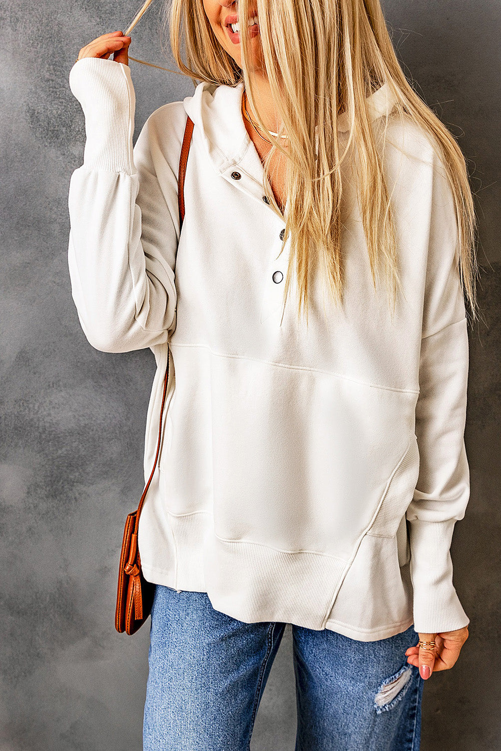 A person with long blonde hair, wearing a White Batwing Sleeve Pocketed Henley Hoodie and blue jeans, stands with their back to the camera, holding a small brown bag over their shoulder. The hoodie features a subtle patchwork design that adds an interesting texture to the outfit.