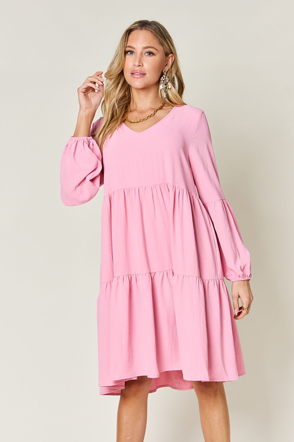 A woman stands wearing the Double Take Full Size V-Neck Balloon Sleeve Tiered Dress with Pockets in pink. Crafted from high stretch yarn, this dress combines comfort and style effortlessly. She has long, wavy hair and accessorizes with hoop earrings and a necklace. This imported garment is easy to care for – simply machine wash cold to keep it looking fresh.