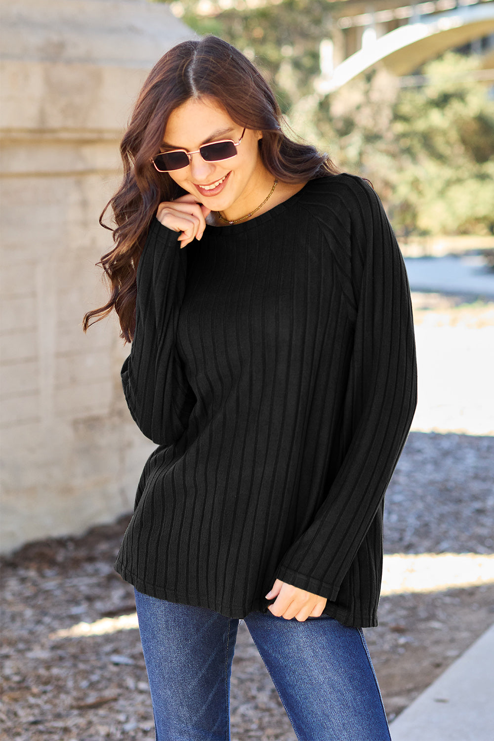 A woman with long brown hair wearing sunglasses and a Basic Bae full-size, slightly stretchy ribbed round neck long sleeve knit top in red, paired with blue jeans, is standing outdoors.