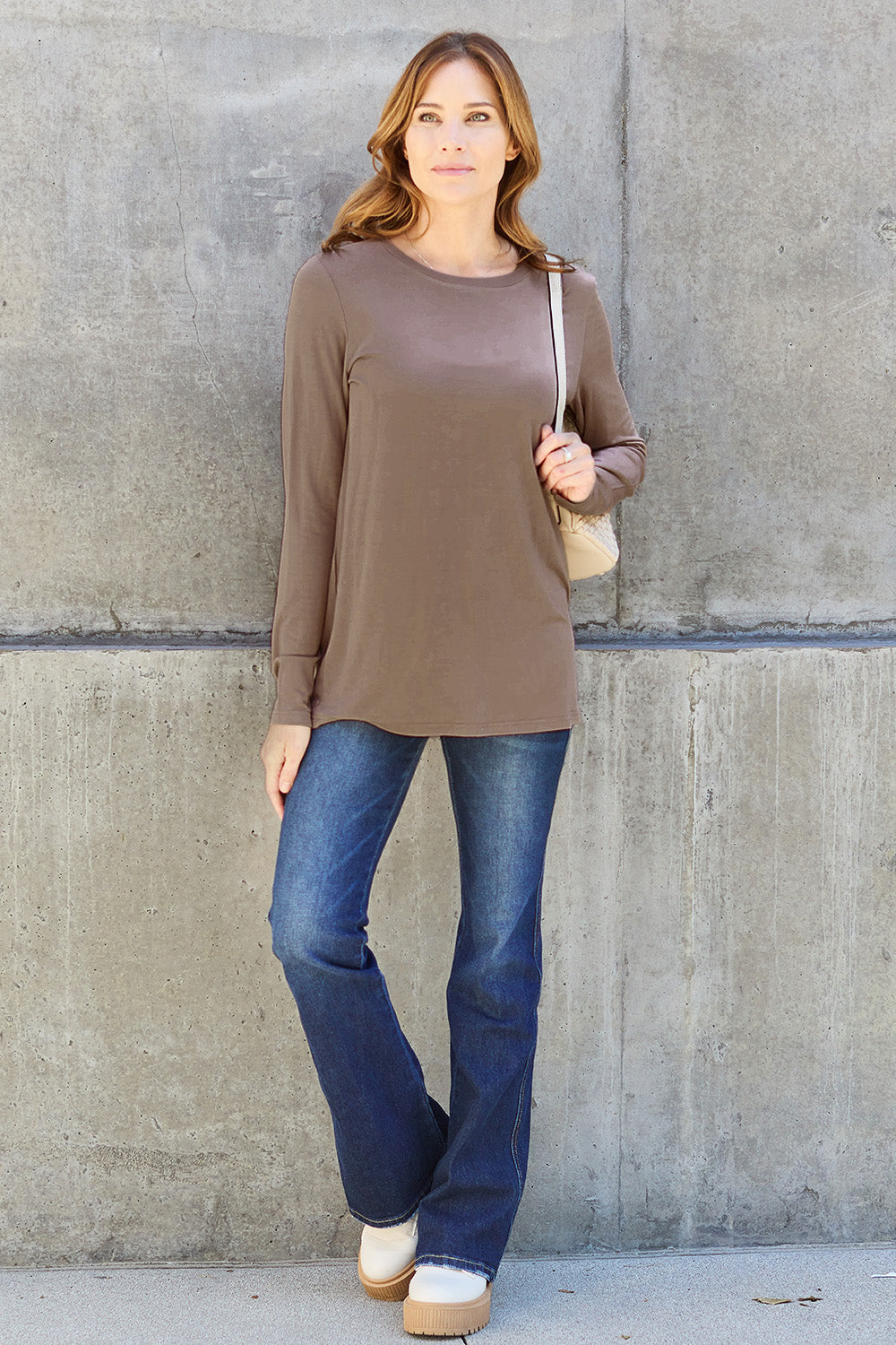 A woman with shoulder-length hair wearing the Basic Bae Full Size Round Neck Long Sleeve Top and blue jeans stands in front of a concrete wall, holding a beige handbag over her shoulder. Her outfit exemplifies basic style, with the top made from 100% polyester and recommended for machine wash cold.