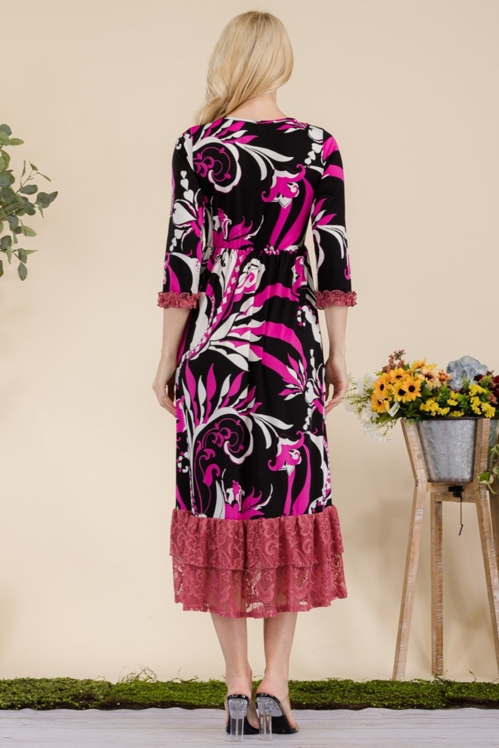 A woman wearing the Celeste Full Size Paisley Print Lace Ruffled Midi Dress poses gracefully for the camera indoors, standing beside metal planters filled with flowers.