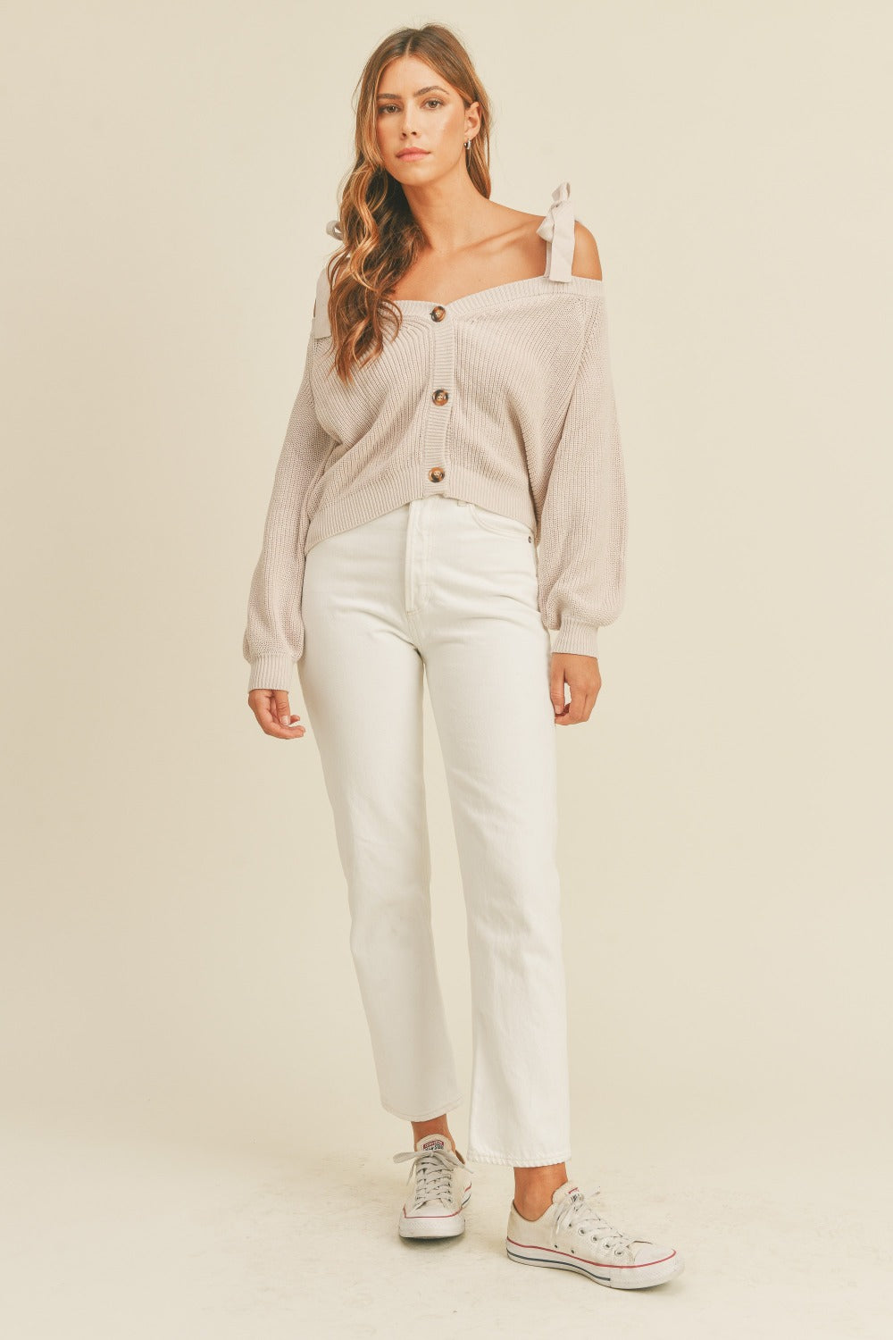 A woman with long, wavy hair is wearing a MABLE Off Shoulder Self Tie Strap Button Down Sweater in beige, paired with white pants, standing against a plain background.