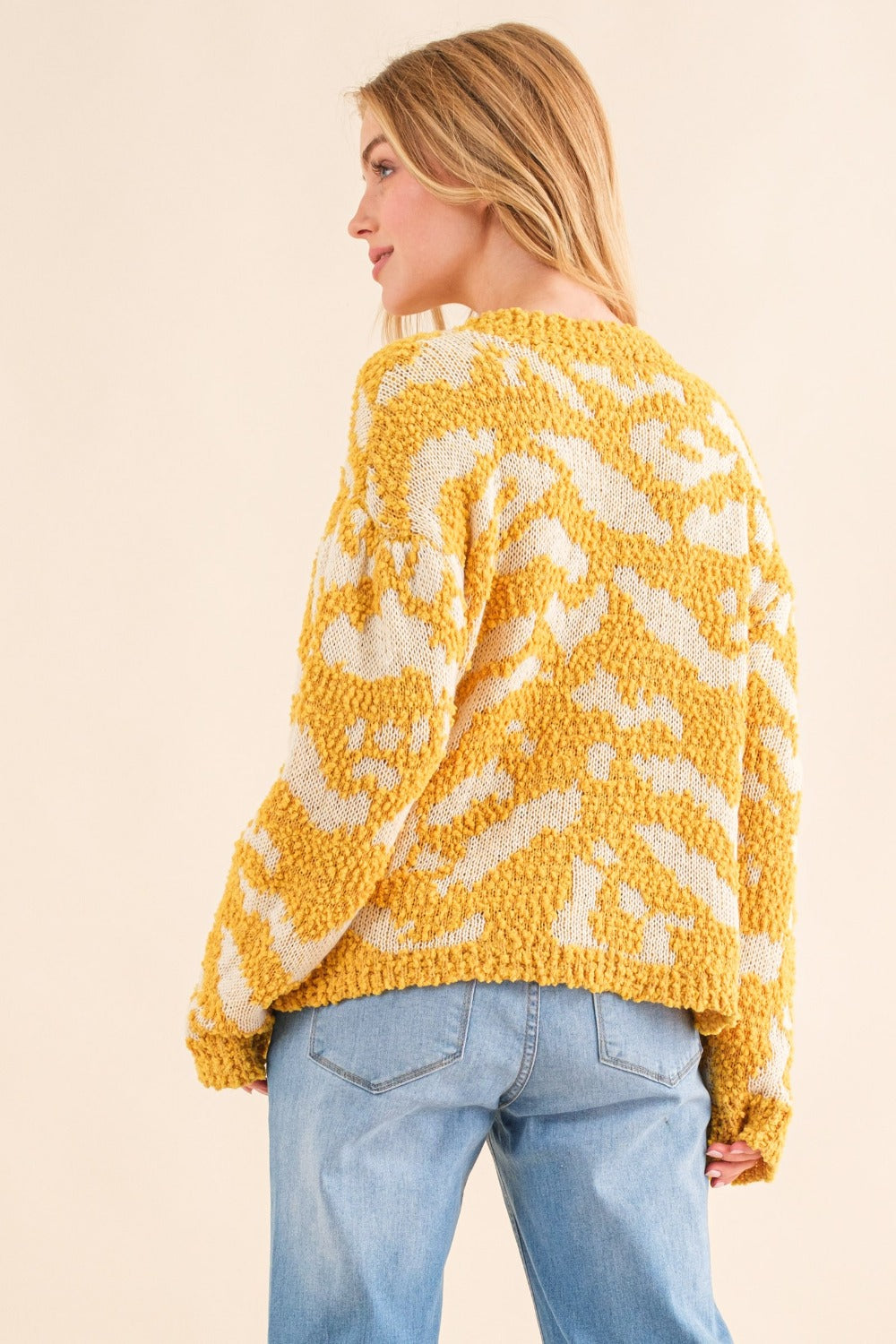 A person with long hair wearing a stylish yellow and white And The Why Full Size Textured Pattern Contrast Sweater and blue jeans stands against a beige background.