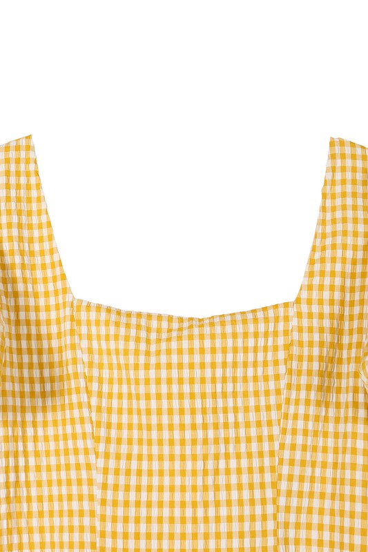 A woman stands indoors, smiling while wearing the SS back strap dress in yellow gingham, and holding a white purse.