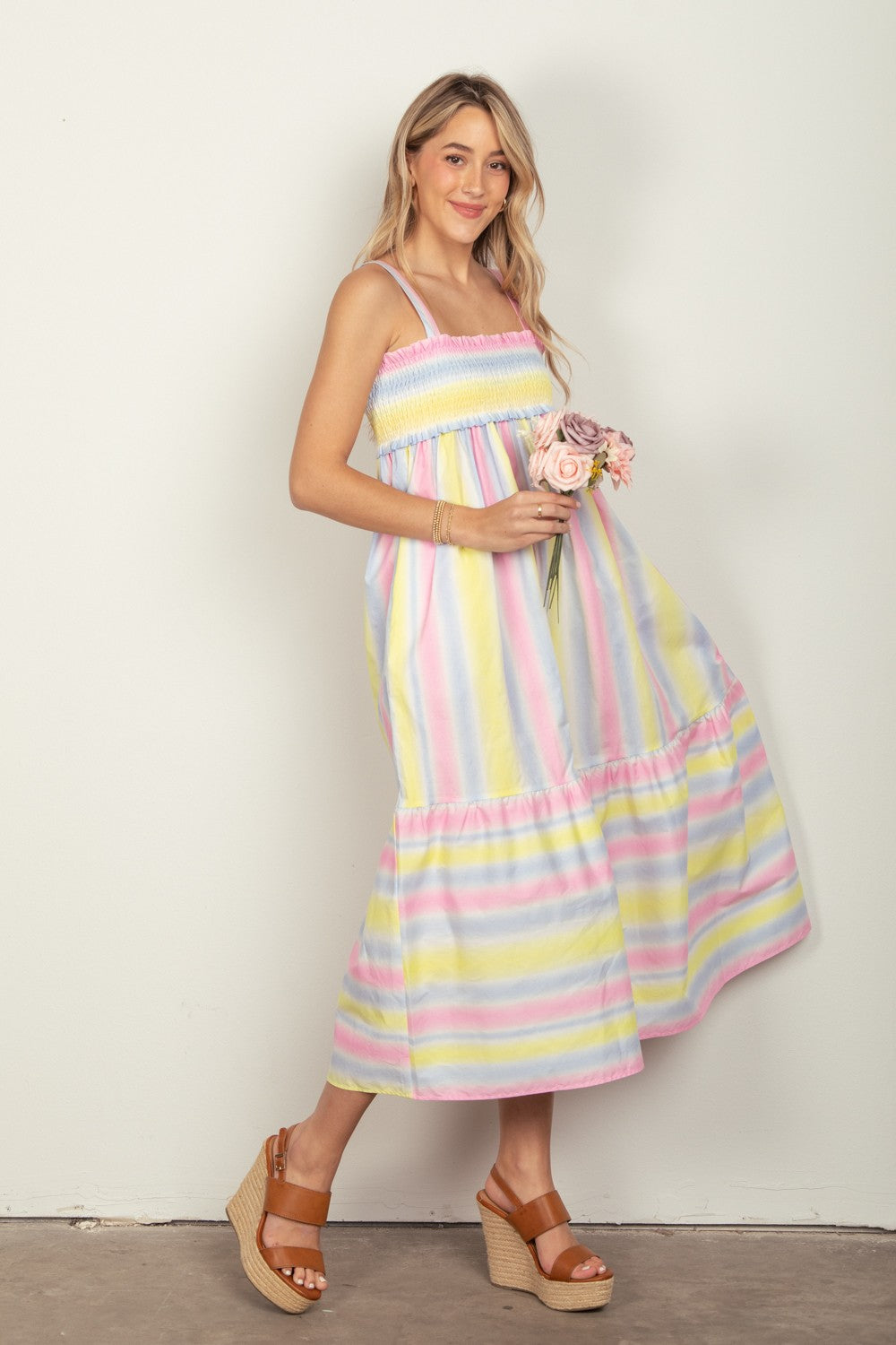 A woman in the VERY J Striped Woven Smocked Midi Cami Dress, featuring pastel stripes, holds a small bouquet of flowers. She pairs the dress with tan platform sandals that complete her chic summer look and stands against a plain white background.