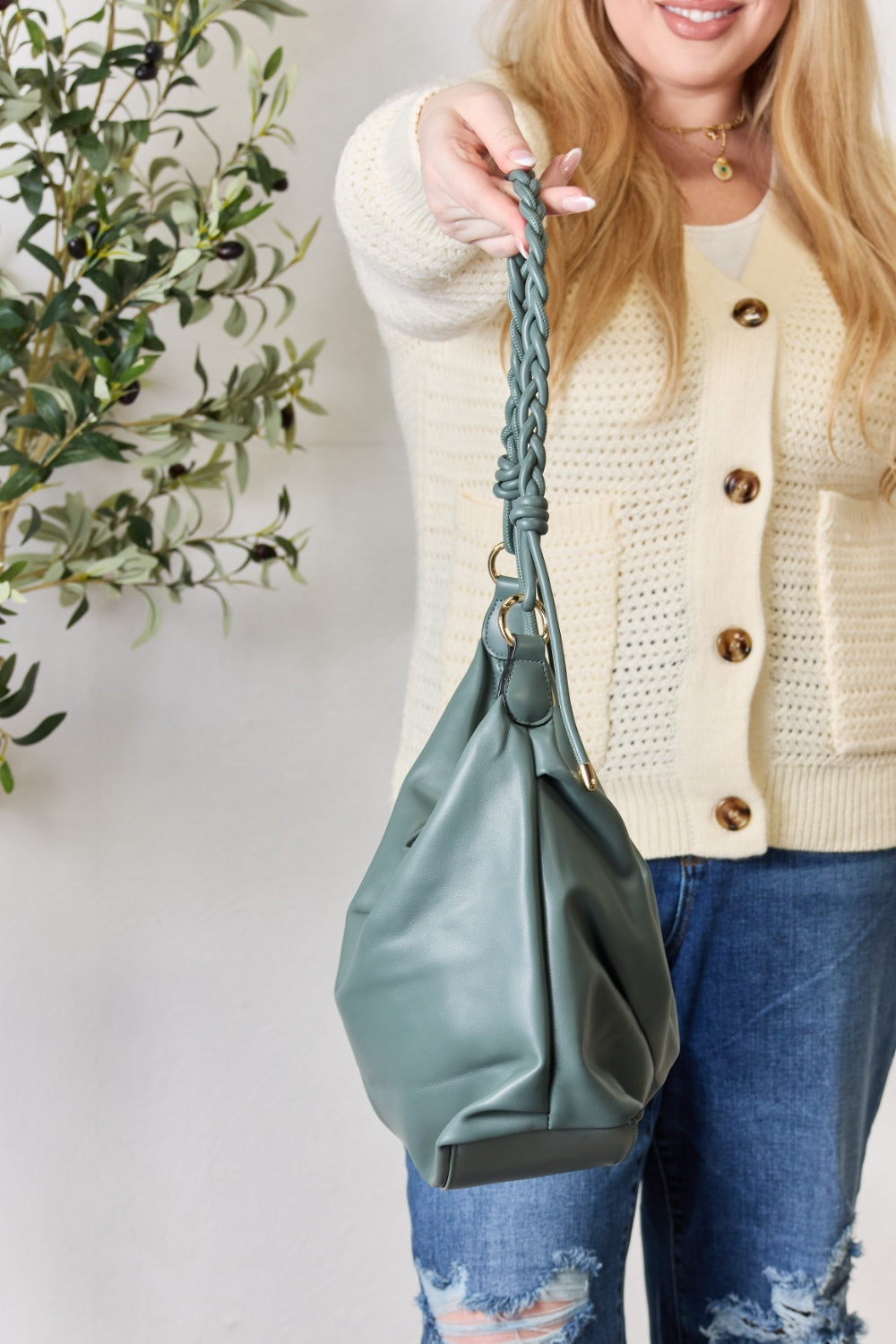 A person in a knitted sweater and jeans holds the SHOMICO Braided Strap Shoulder Bag, a blue-green statement accessory featuring straps crafted from vegan leather.