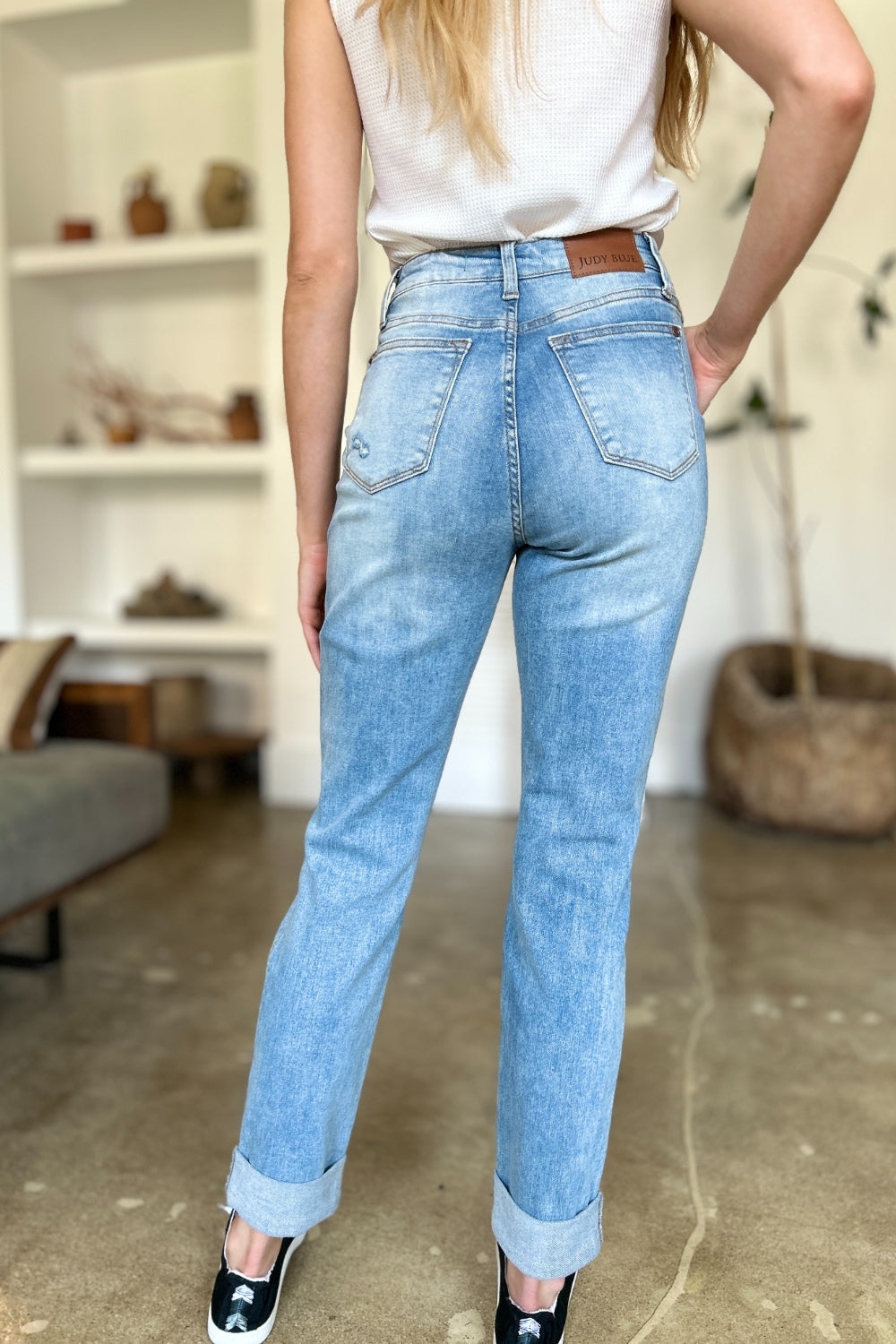 Two women are standing side by side, both clad in black and white tank tops and Judy Blue Full Size Distressed Straight Jeans with Patch Pockets, smiling at the camera in a modern living room.