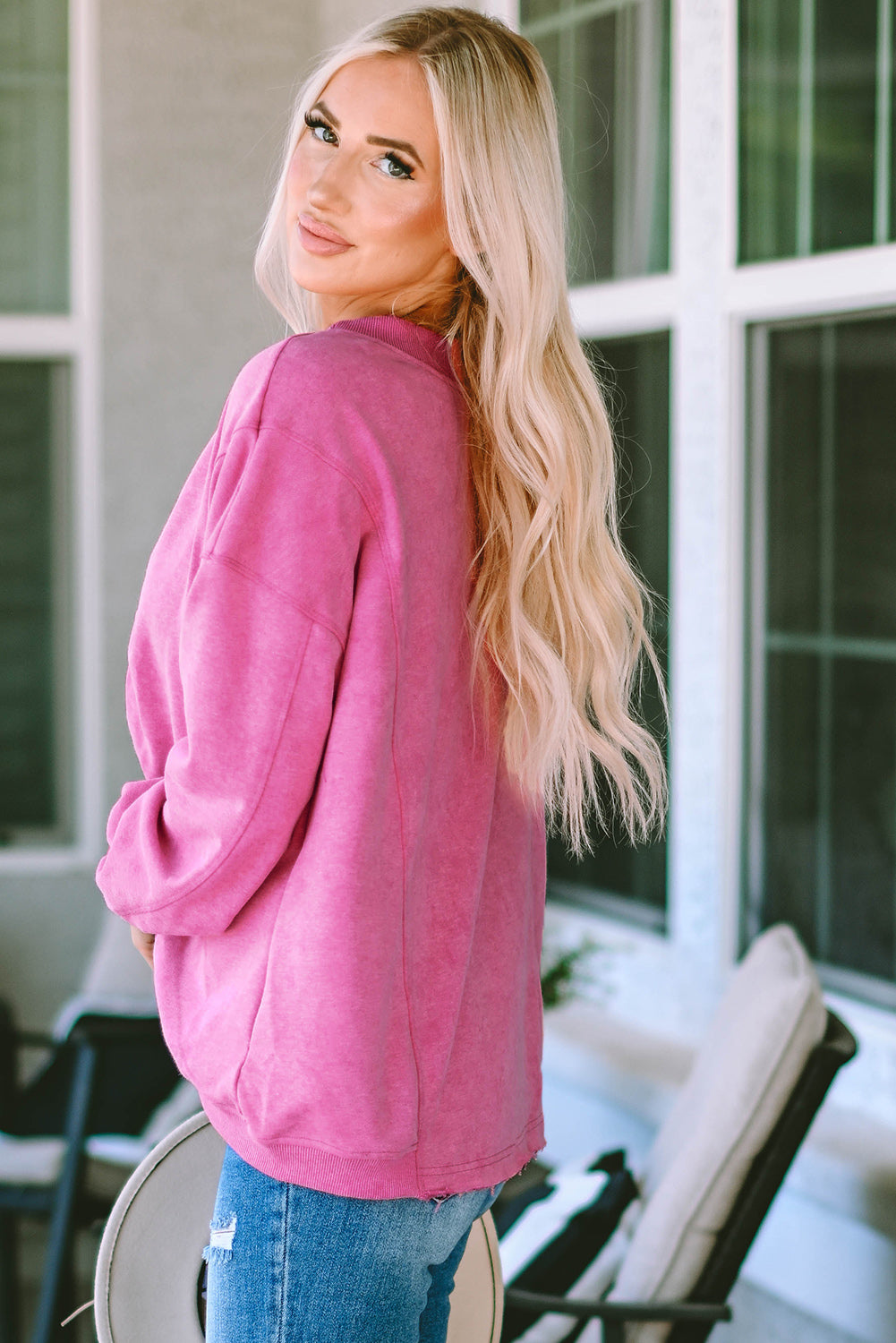 A woman stands with her back to the camera, wearing a long-sleeved Rose Exposed Seam Twist Open Back Oversized Sweatshirt and light blue jeans, showcasing a relaxed fit.