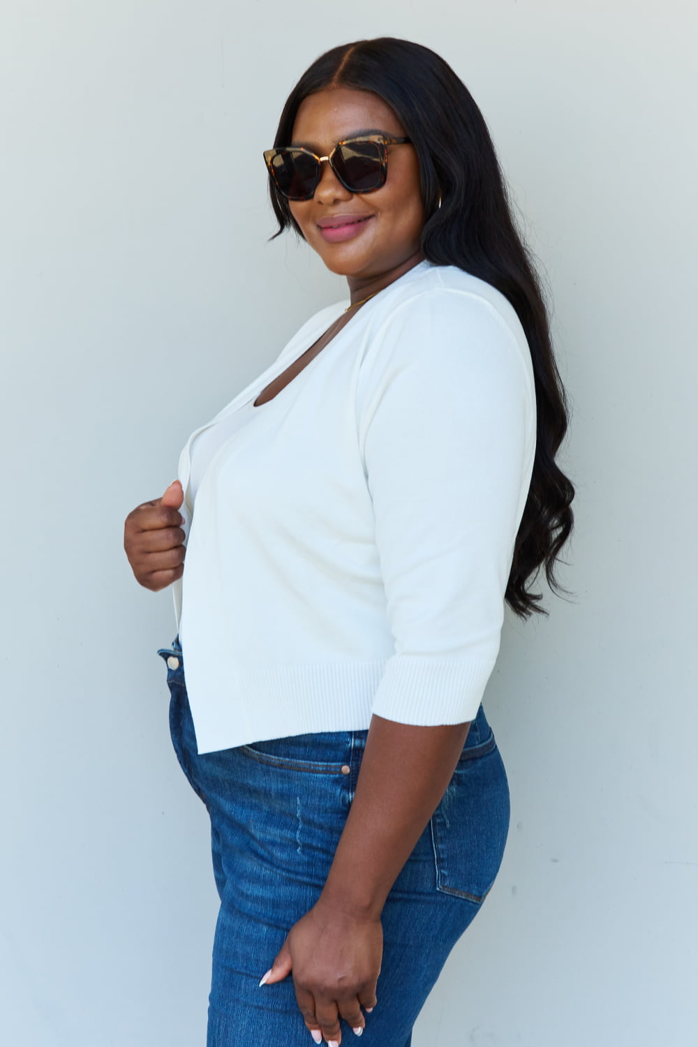 A stylish person wearing sunglasses, a Doublju My Favorite Full Size 3/4 Sleeve Cropped Cardigan in Ivory, and blue jeans poses in front of a plain background, effortlessly highlighting their versatile look.