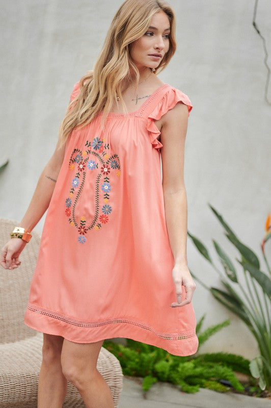 A woman in an Embroidered Square Neck Mini Dress, featuring a coral hue, stands near plants adjusting her hair—the perfect summer attire.