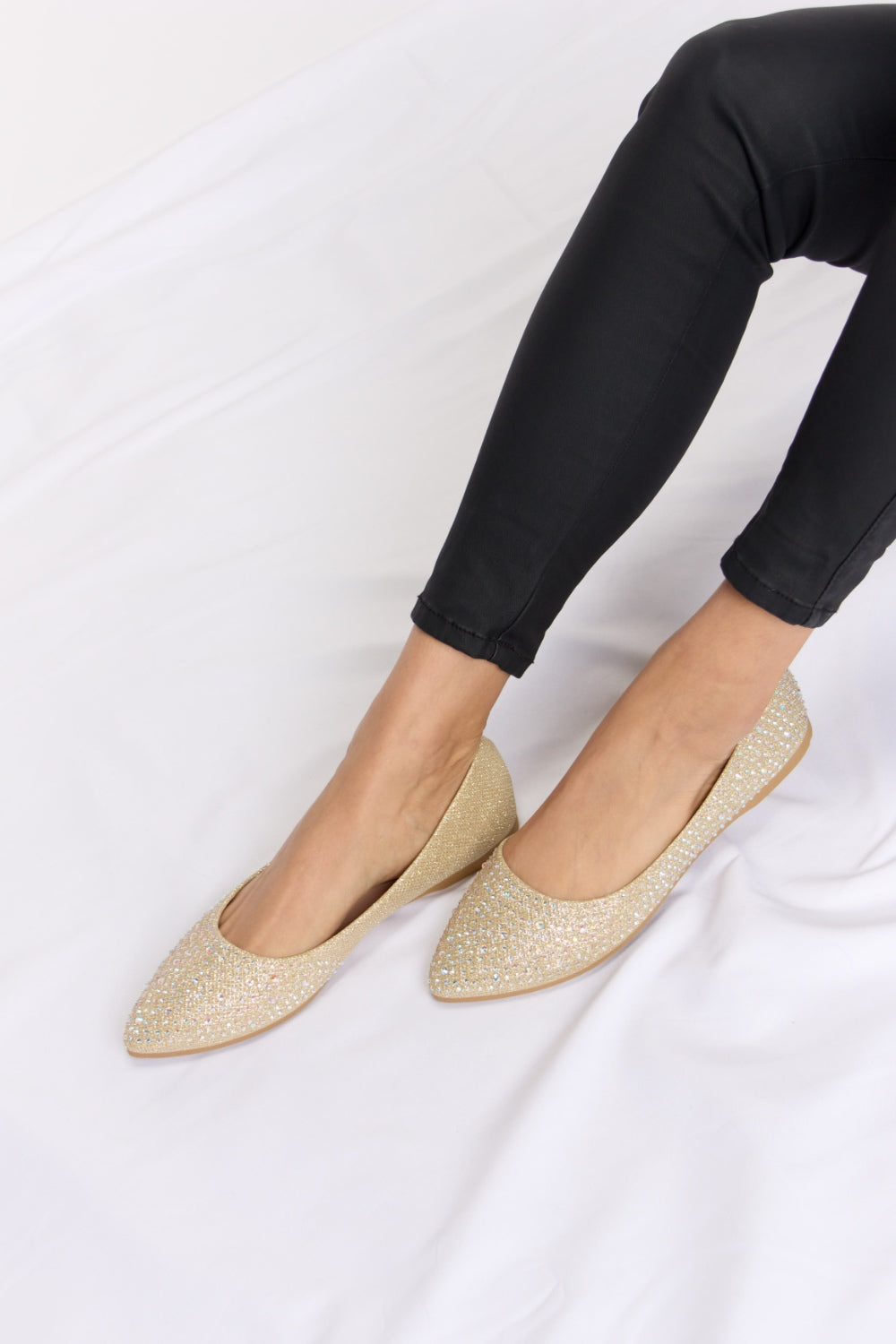 Someone is sitting on a white surface wearing black pants and chic Forever Link Rhinestone Point Toe Flat Slip-Ons in sparkling gold.