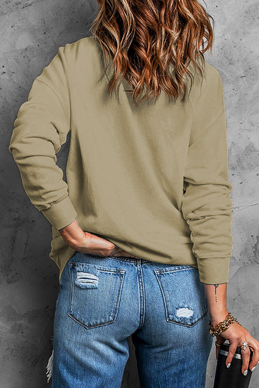 A person with wavy hair, wearing a Khaki Plain Crew Neck Pullover Sweatshirt and ripped blue jeans, stands with their back to the camera against a grey concrete wall.