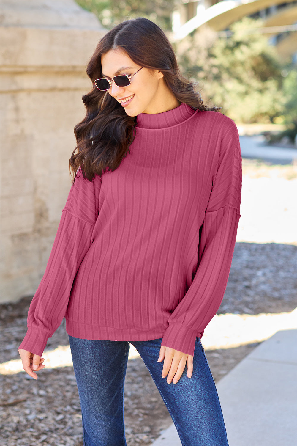 A woman with long hair is wearing a green Basic Bae Full Size Ribbed Exposed Seam Mock Neck Knit Top, blue jeans, and sunglasses. She is standing outdoors and smiling.