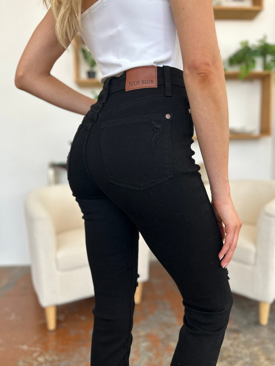 A person wearing a white T-shirt and Judy Blue Full Size Distressed Tummy Control High Waist Skinny Jeans poses barefoot in a room with white chairs and decorative plants in the background.