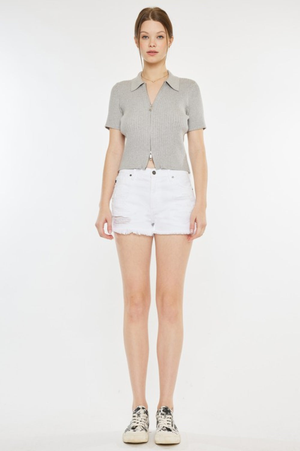 A person stands against a plain background, wearing a light grey short-sleeve top, Kancan Raw Hem Distressed Denim Shorts in white, and white slip-on shoes with black patterns.