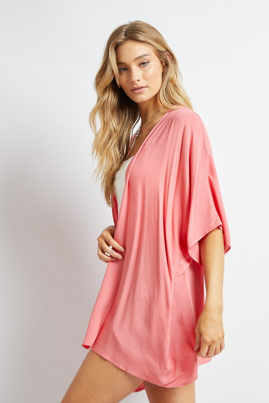 A woman poses against a simple backdrop, gracefully wearing a wardrobe essential: the Favorite Solid Kimono Cardigan in pink. Complementing her white tank top and denim shorts, this casual layer enhances her relaxed style with an elegant touch.