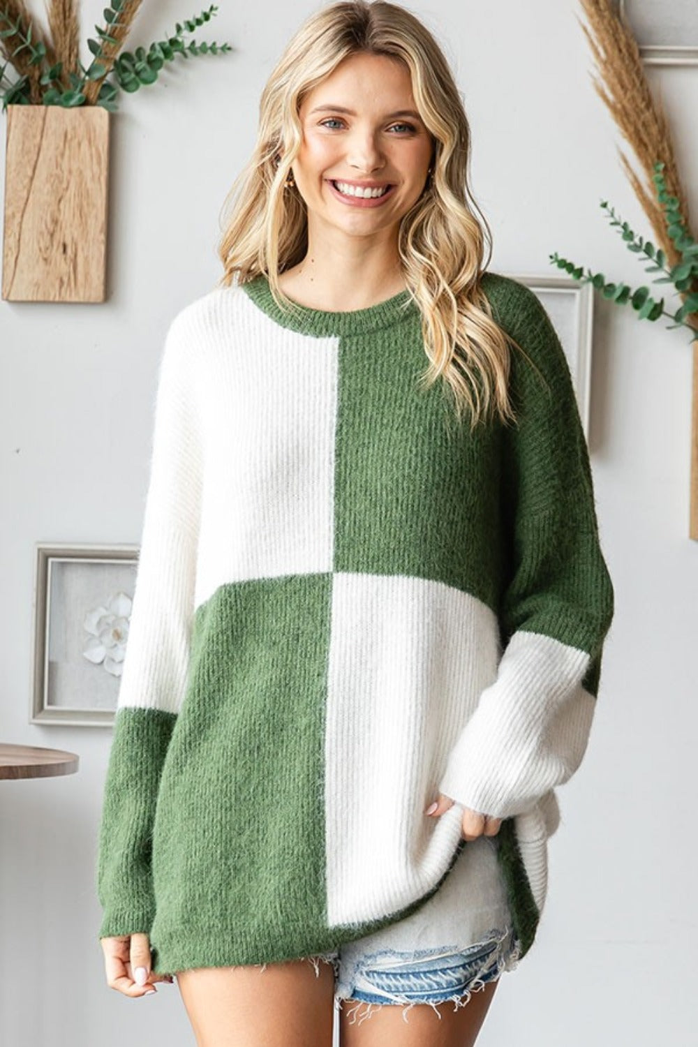 A woman smiles while wearing the First Love Color Block Round Neck Long Sleeve Ribbed Sweater in green and white, paired with denim shorts and a wide-brimmed hat. She is sitting on a chair with plants in the background.