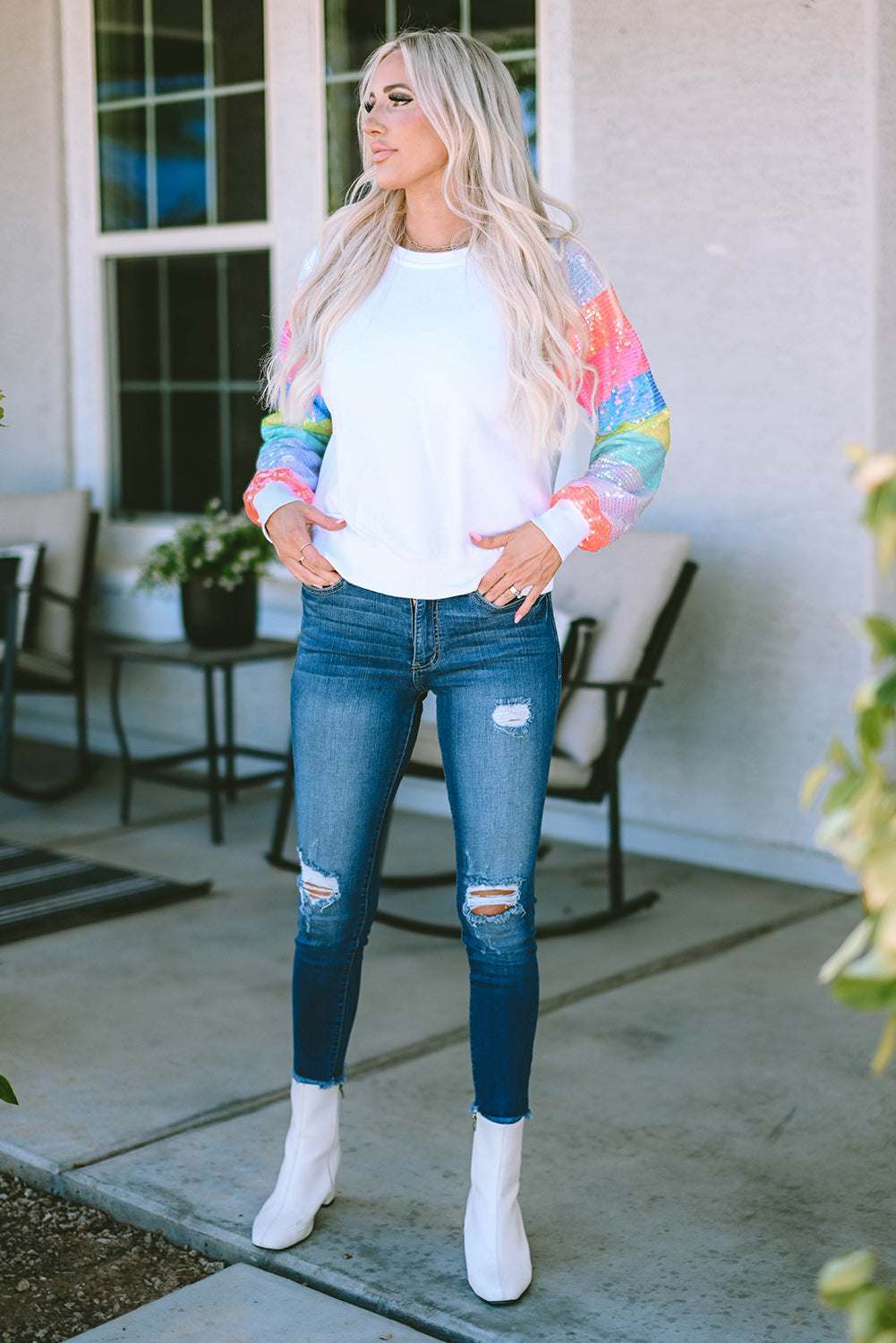 A person with long blonde hair is seen from the back, wearing a White Sequin Color Block Raglan Sleeve Pullover Sweatshirt and blue jeans. They are standing in front of a building with large windows.