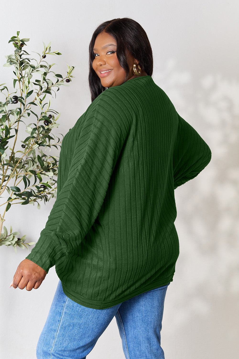 A person with wavy hair, wearing the Basic Bae Full Size Ribbed Cocoon Cardigan over a white top and beige pants, stands in front of a light-colored background with a leafy plant, showcasing an effortlessly basic style.