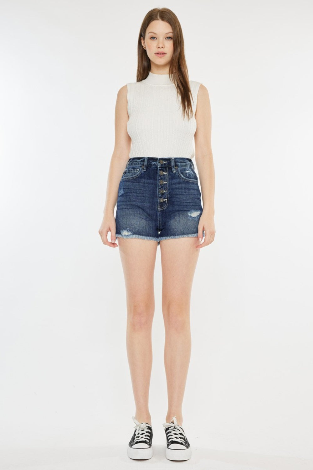 A woman stands against a plain background wearing a sleeveless white top, Kancan Raw Hem Button Fly Denim Shorts, and black slip-on sneakers.