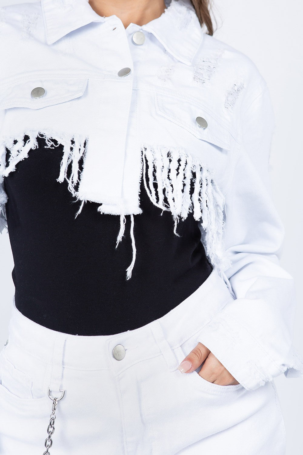 A woman in an American Bazi Distressed Denim Jacket with Frayed Hem over a black top, paired with frayed white high-waisted shorts, stands with one hand in her pocket against a white background.