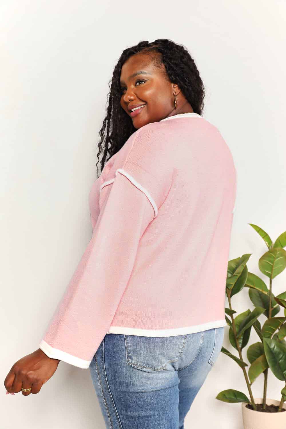 A woman with long curly hair is wearing the Angel Wings Contrast Detail Dropped Shoulder Knit Top in pink, paired with blue jeans, standing against a light background.