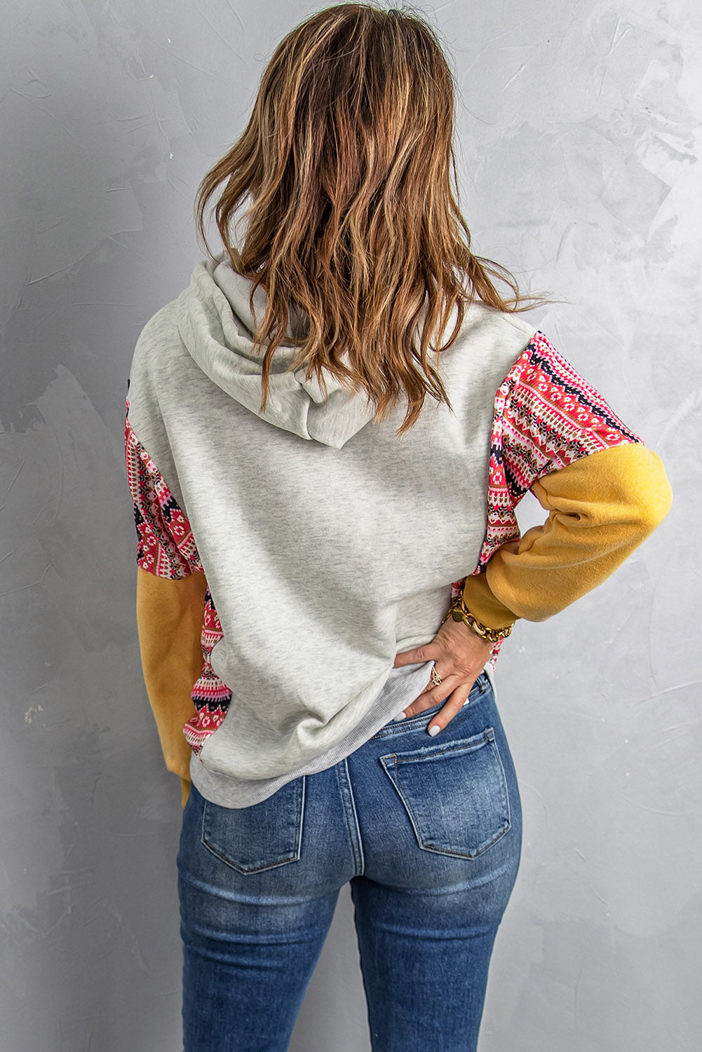 A person with long hair wearing an Aztec Patch Pullover Hoodie and blue jeans is facing away from the camera against a textured gray wall.