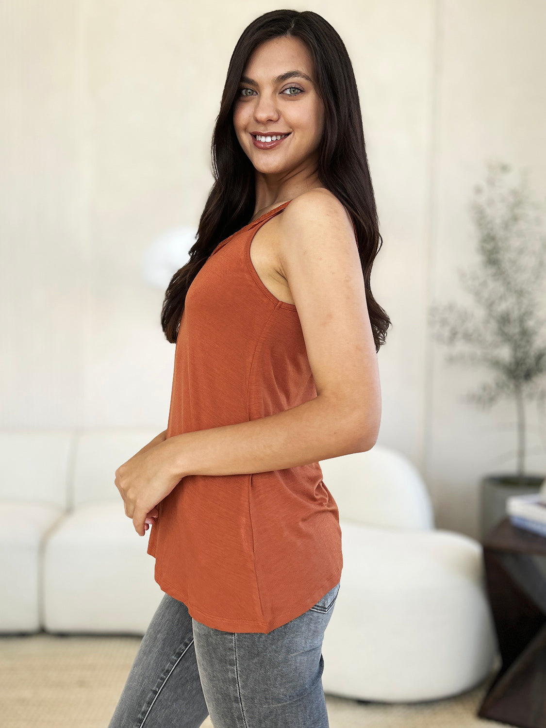 A woman with long hair wearing sunglasses, a slightly stretchy pink "Basic Bae Full Size Lace Detail V-Neck Cutout Cami," and basic style blue jeans stands against a concrete wall, looking to the side with one hand touching her upper chest.