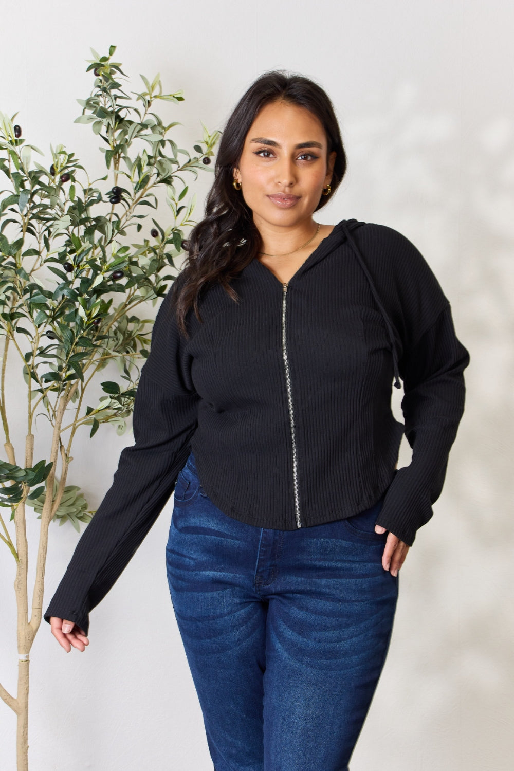 A person wearing the Culture Code Full Size Ribbed Zip Up Drawstring Hooded Jacket and blue jeans stands in front of a plain background with a small leafy plant to the left.