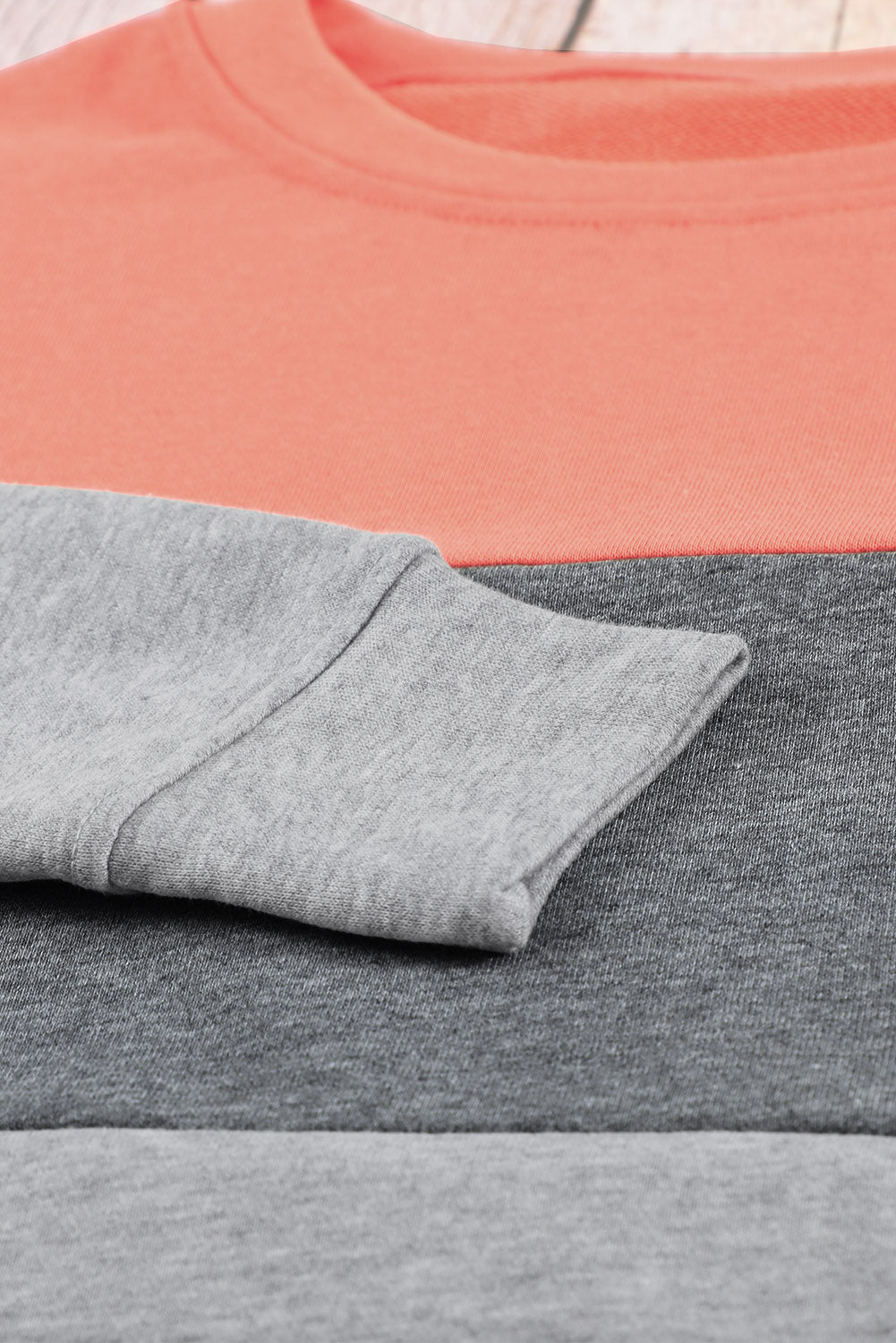 A Colorblock Gray Contrast Stitching Sweatshirt with Slits, made of comfy fabric and featuring coral, gray, and light gray sections, is placed on a wooden surface near sunglasses, a book, and some plant leaves.