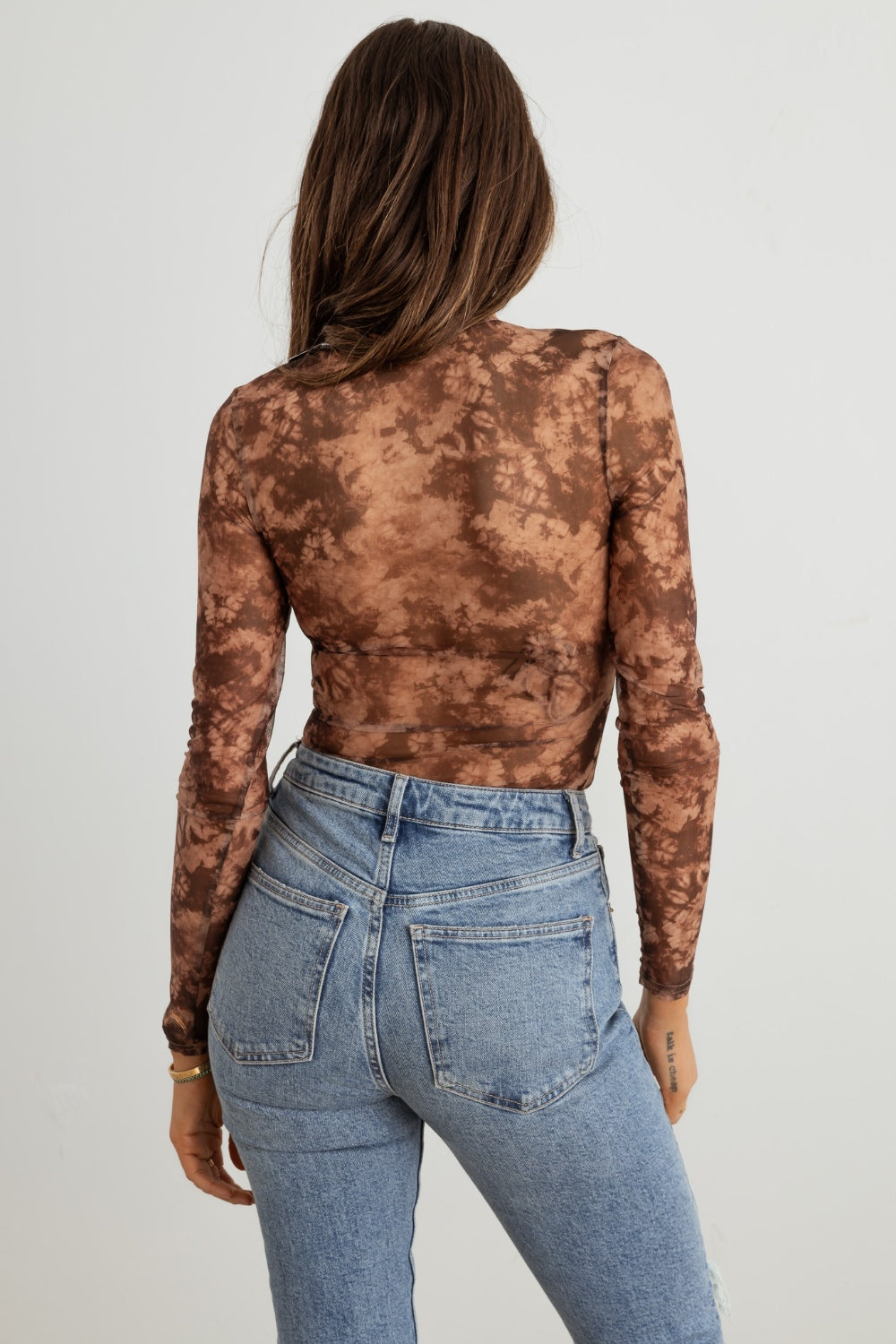 A woman with long hair is wearing the HERA COLLECTION Abstract Mesh Lace-Up Long Sleeve Bodysuit paired with ripped blue jeans, creating a statement-making look as she stands against a plain background.