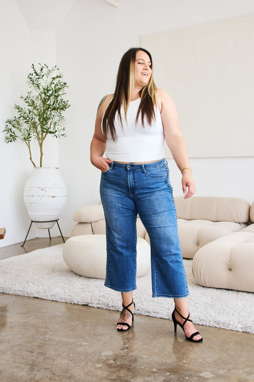 A person wearing Judy Blue Full Size Braid Side Detail Wide Leg Jeans and black heels is standing on a polished floor. There is a white couch and a large potted plant in the background.