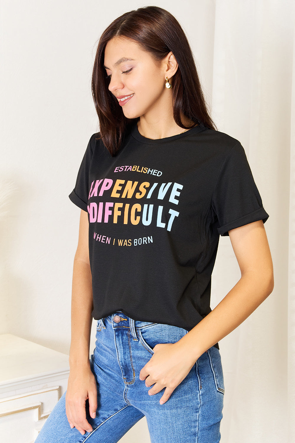 A woman with long braided hair, wearing the Simply Love Slogan Graphic Cuffed Sleeve T-Shirt that says "Established Expensive & Difficult When I Was Born," stands with one hand in her pocket. The trendy cuffed sleeves of her shirt add an extra touch of style.