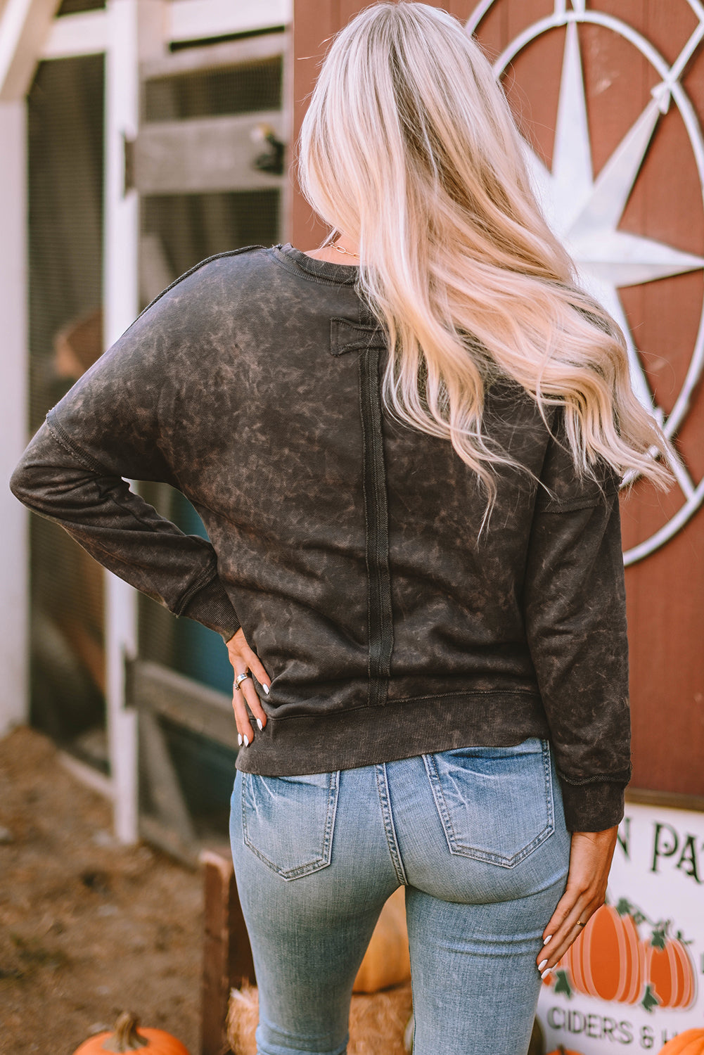 A person with long blonde hair stands with their back to the camera, wearing a Gray Acid Wash Drop Shoulder Long Sleeve Sweatshirt with Pockets and jeans. A large star decoration is visible in the background, along with some pumpkins on the ground.