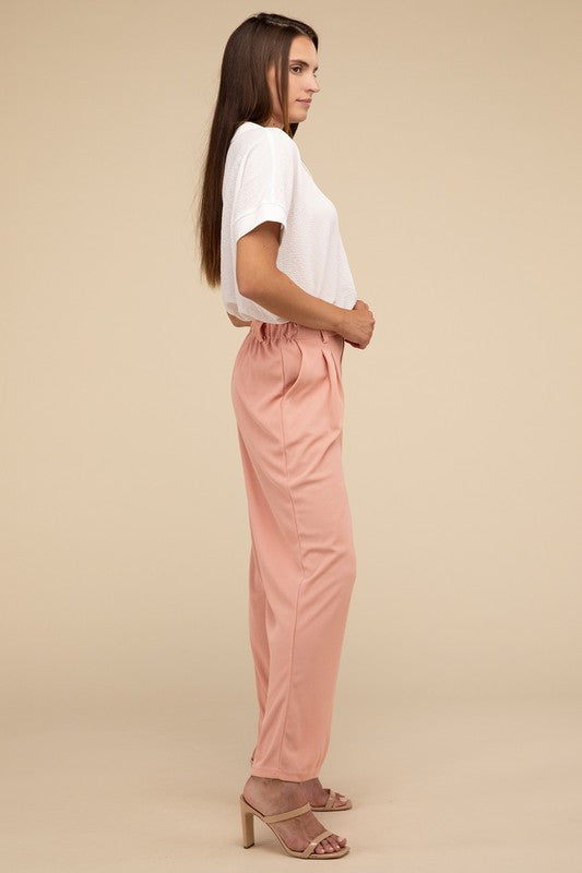 A woman stands against a beige background, highlighting her chic ensemble with a tailored fit. She wears a white short-sleeve shirt paired with versatile pink Waffle Trouser Pants, hands casually in pockets, complemented by stylish strappy sandals.