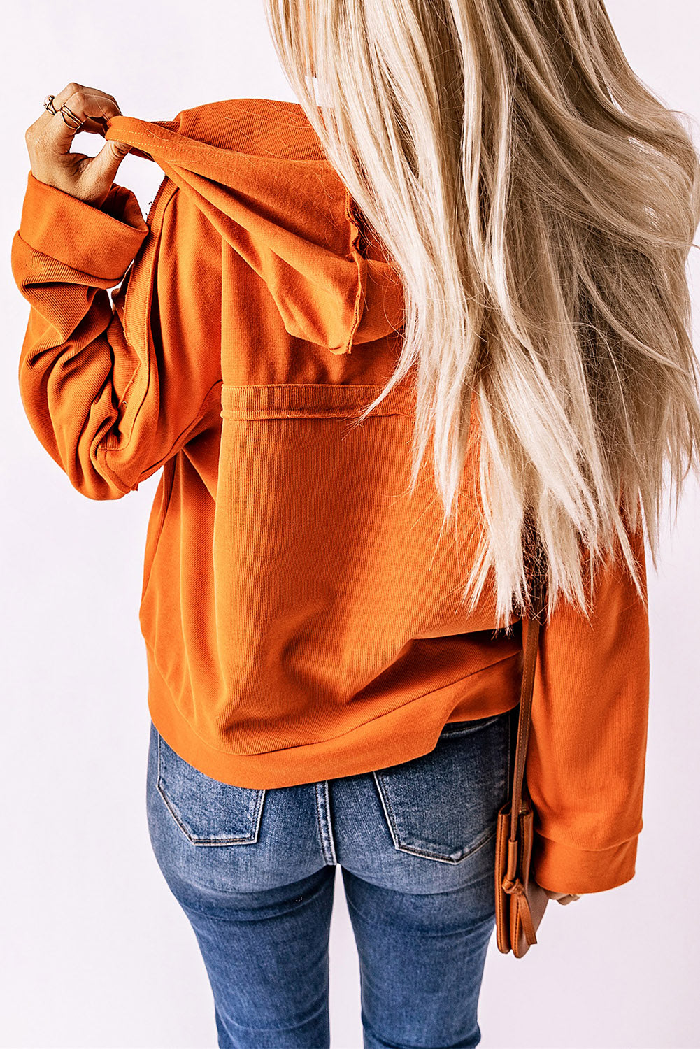 A person with long blonde hair, dressed in the Orange Casual Button Solid Patchwork Trim Hoodie and blue jeans, is holding the hood of their hoodie with one hand while facing away from the camera. This outfit is ideal for fall and winter essentials.